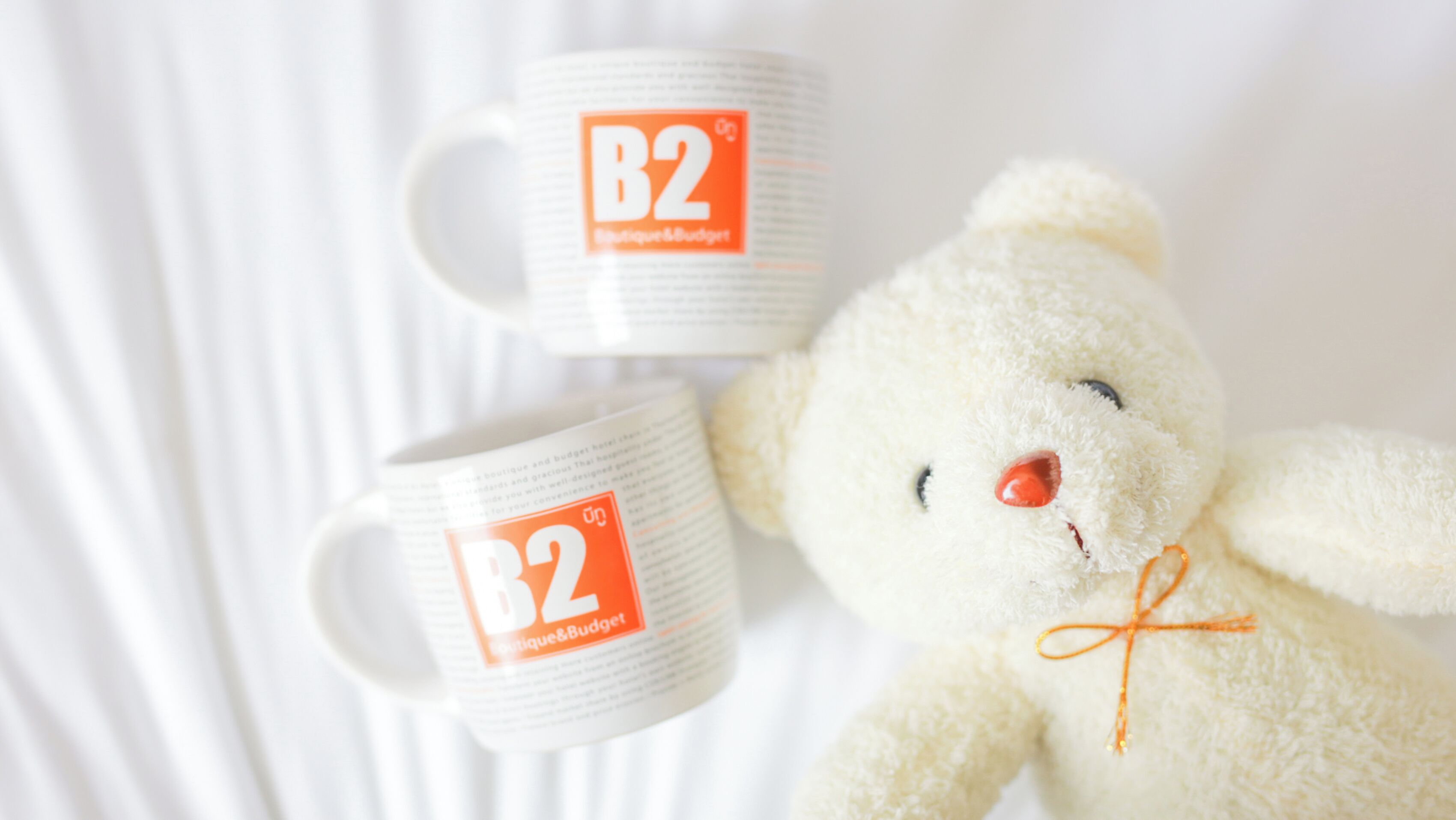 Een witte knuffelbeer met een rode neus en vlinderdas, liggend naast twee witte mokken met oranje B2 logo's.