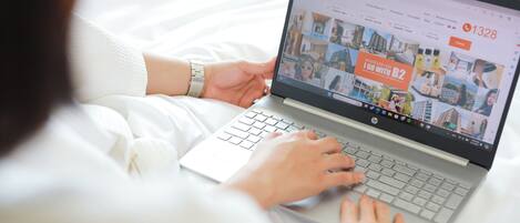 Una laptop sobre una cama mostrando un sitio web de reservas de hotel con varias habitaciones y un número de contacto.