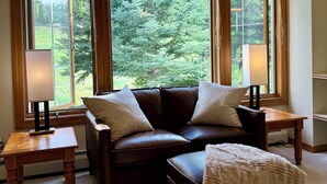 A cozy living room with a dark brown leather sofa, two matching armchairs, a wooden coffee table, and a white throw blanket on a beige carpet. Large windows with wooden frames offer a view of a green landscape. - Large Cozy Townhouse Ski-in / Ski-out to Super Bee, Private Washer /Dry & Garage (Frisco)