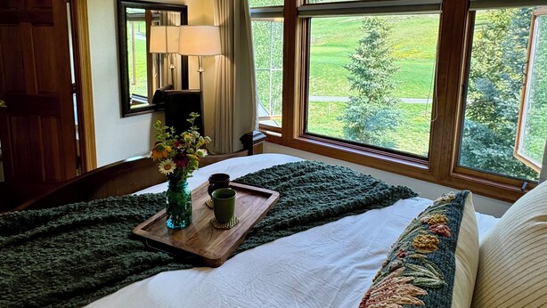A bedroom with a large window, a wooden bedside table with a vase and two cups, a green knitted throw, and a bed with a white comforter and a colorful pillow. - Large Cozy Townhouse Ski-in / Ski-out to Super Bee, Private Washer /Dry & Garage (Frisco)