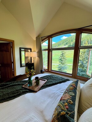 A bedroom with a large window, a wooden bedside table with a vase and two cups, a green knitted throw, and a bed with a white comforter and a colorful pillow. - Large Cozy Townhouse Ski-in / Ski-out to Super Bee, Private Washer /Dry & Garage (Frisco)