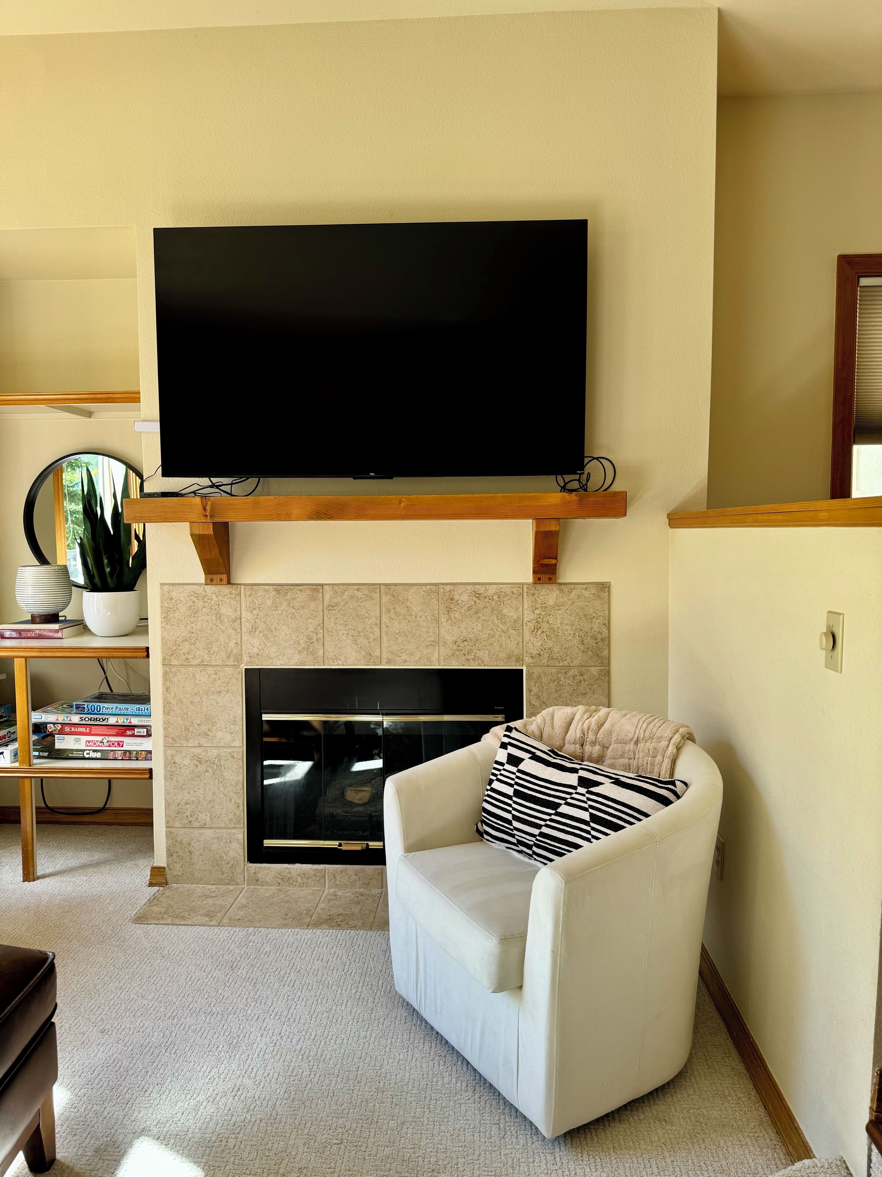 A living room with a flat-screen TV mounted above a fireplace, a white armchair with a striped pillow, and a bookshelf with various items.