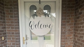 Une porte d’entrée blanche, avec une pancarte « Bienvenue » au-dessus et un paillasson en dessous.