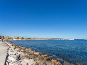 A coastal walkway with a rocky shoreline, a lighthouse in the distance, and buildings along the coast.
