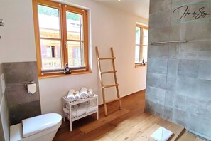 Une salle de bain comprenant des toilettes, une étagère avec des serviettes, une échelle en bois et un espace douche avec un mur carrelé.