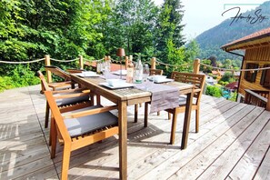 Une terrasse en bois avec une table dressée pour le repas, entourée de chaises, d’où l’on peut admirer un paysage verdoyant et, au loin, une montagne.