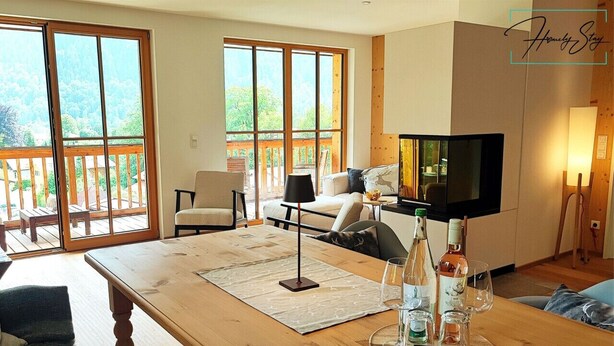 Un grand salon comprenant une table en bois, un canapé et un téléviseur. Il y a également un balcon doté d’une balustrade en bois et offrant une vue sur la verdure.