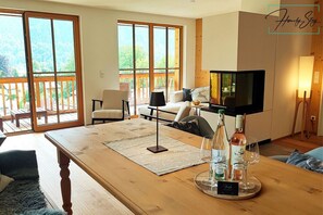 Un grand salon comprenant une table en bois, un canapé et un téléviseur. Il y a également un balcon doté d’une balustrade en bois et offrant une vue sur la verdure.