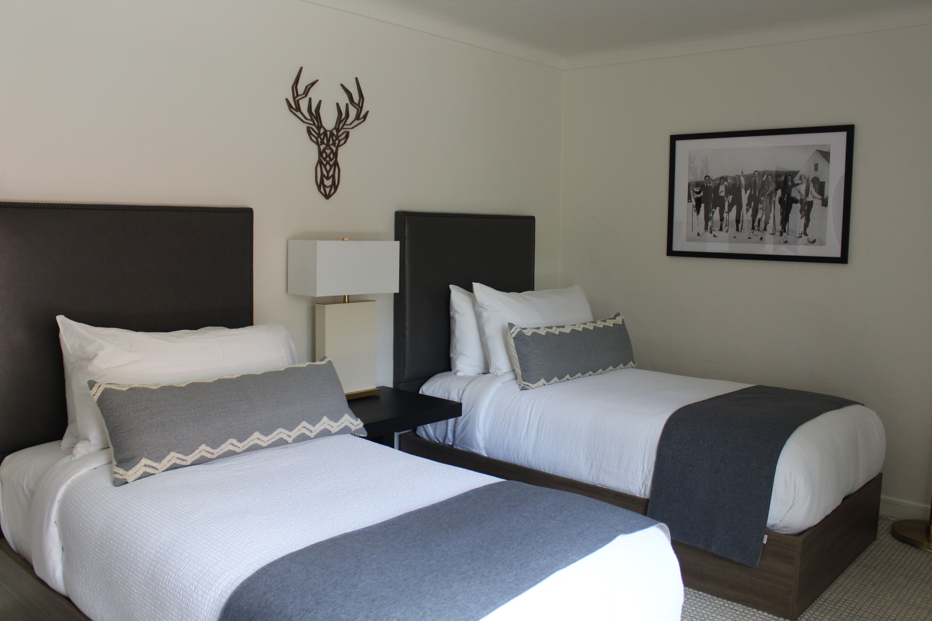 Habitación de hotel con dos camas, cabecero decorado con cabeza de ciervo, fotografía enmarcada en la pared y mesita de noche con lámpara.