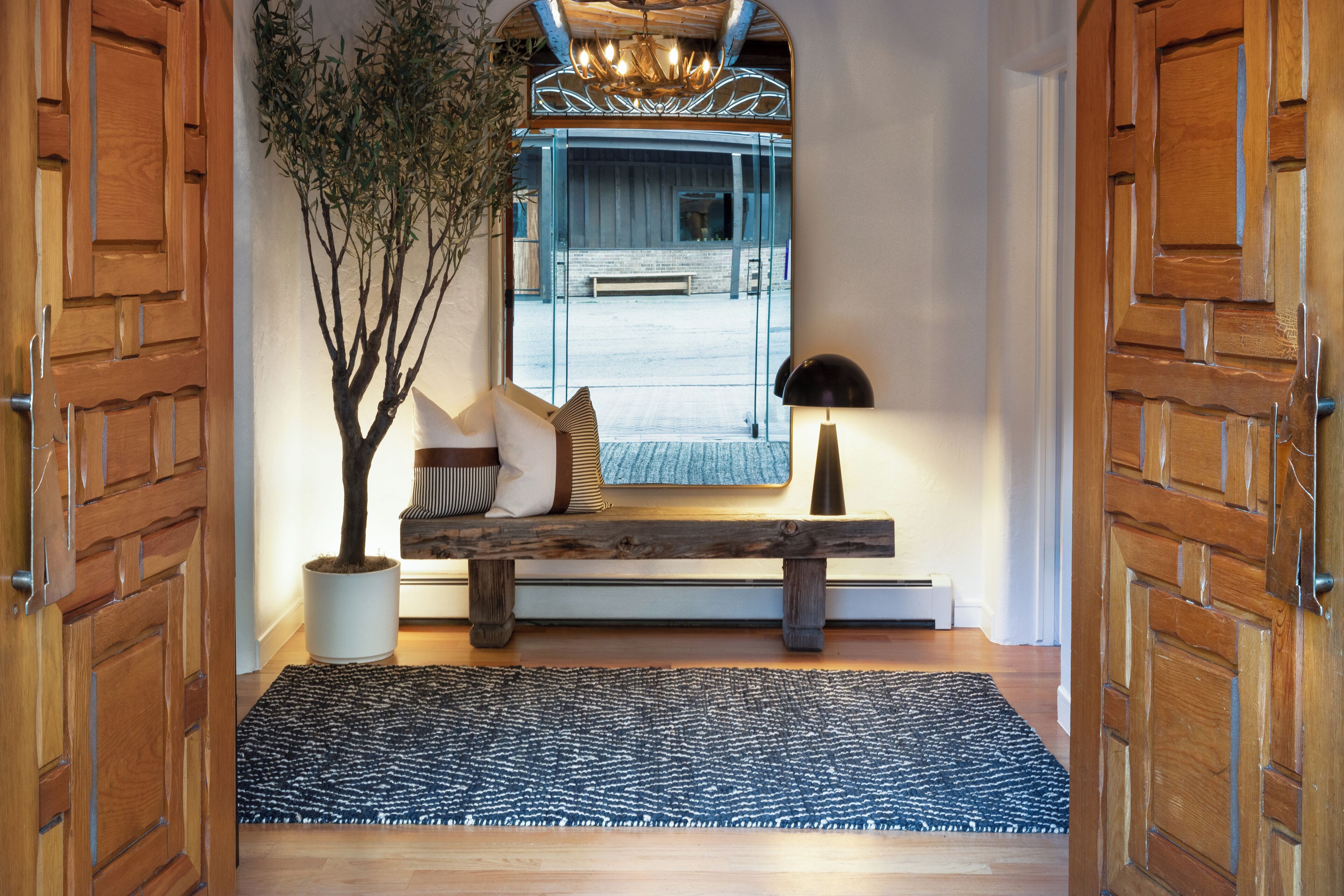 A wooden door with a textured pattern, a bench with cushions, a potted plant, and a rug leading to a room with a bench and a mirror.