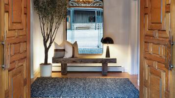 A wooden door with a textured pattern, a bench with cushions, a potted plant, and a rug leading to a room with a bench and a mirror.