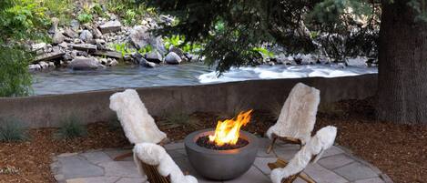 A stone patio with a central fire pit and two fur-covered chairs.