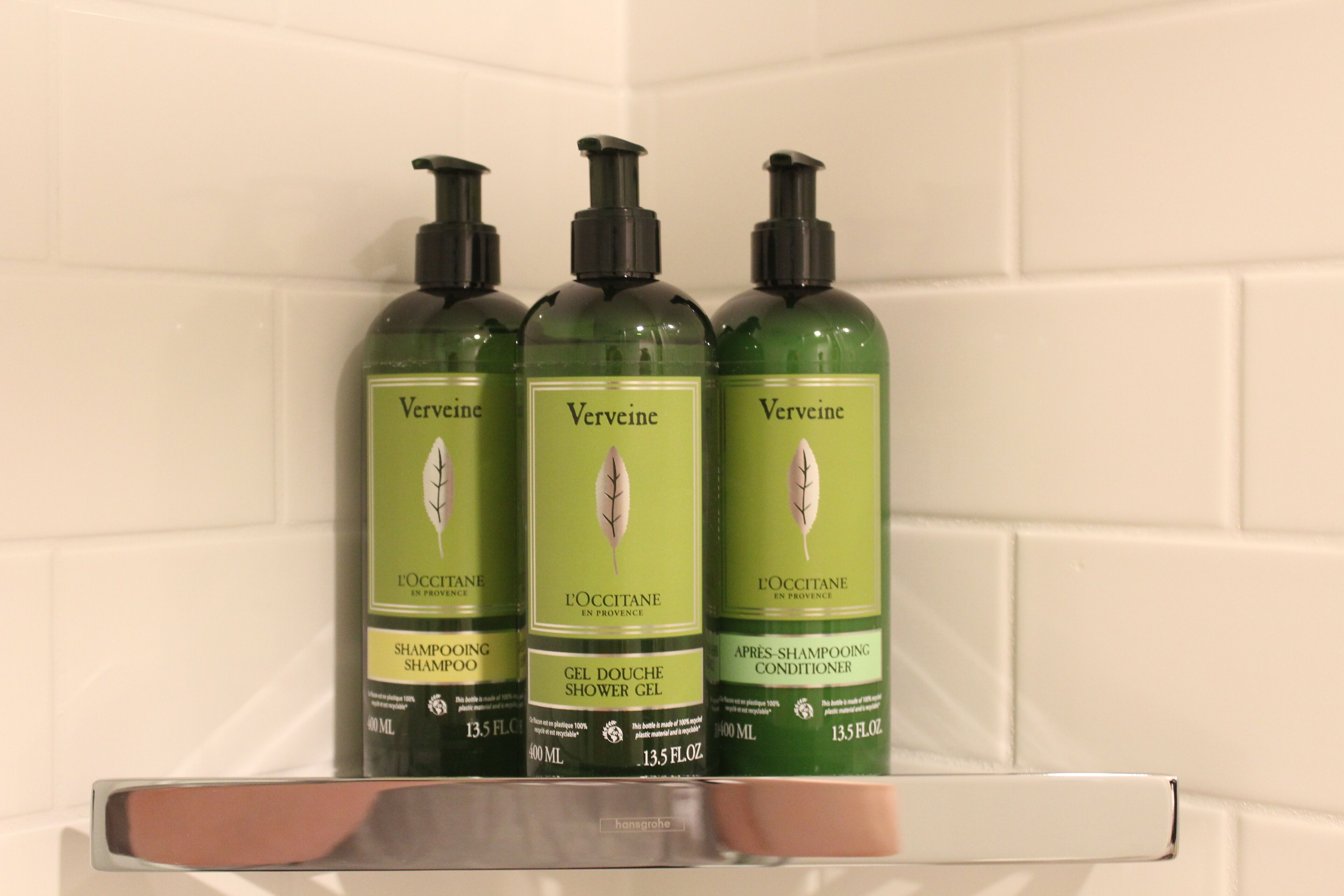 Trois bouteilles de shampoing et d’après-shampoing à la verveine, posées sur une étagère.