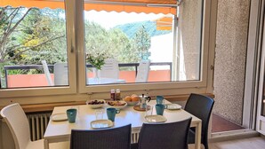 A dining area with a table set for two, a view of greenery and mountains through large windows, and a partial view of a room with a textured wall.