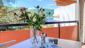A table with a clear vase containing flowers, a book, and two mugs on a balcony with a view of trees and hills.