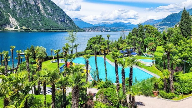 A resort with a swimming pool surrounded by lush greenery and palm trees, overlooking a lake and mountains.
