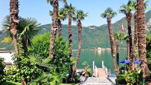 A stone pathway leading to a dock with palm trees and greenery on either side, overlooking a calm lake and a mountainous backdrop.