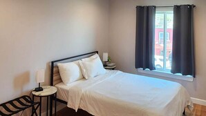 A bedroom with a bed, a bedside table, a lamp, a window with curtains, and a view of a building outside.