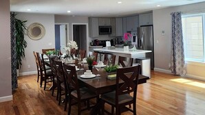 A spacious dining area with a wooden floor, a central dining table set for six, and a kitchen in the background.