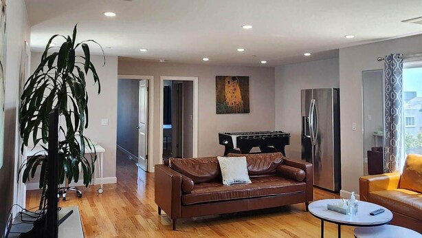 A spacious living room with wooden flooring, a brown leather sofa, a white coffee table, and a large plant.