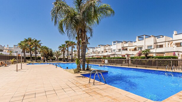 A swimming pool with tiled flooring, palm trees, and modern buildings in the background.