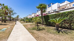 A residential area with modern white buildings, a concrete pathway, and well-maintained greenery including palm trees and hedges.