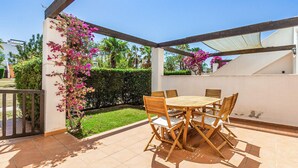 An outdoor patio with a wooden table and chairs, surrounded by a white wall and a garden with pink flowers.