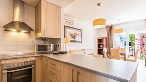 A modern kitchen with wooden cabinets, a built-in oven, and a microwave. There is a dining area with a table and chairs, and a view of the outdoors through a large window.