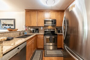 Una cocina con armarios de madera, electrodomésticos de acero inoxidable, incluyendo un refrigerador, microondas y horno, una encimera de granito y una pequeña alfombra.