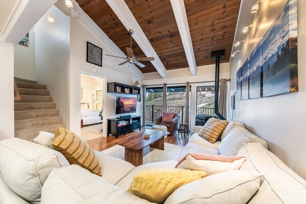A spacious living room with a wooden ceiling, a television, a fireplace, and a balcony with a scenic view.