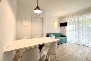 A modern living room with a white dining table, white chairs, a teal sofa, a mounted TV, and a large window with sheer curtains.