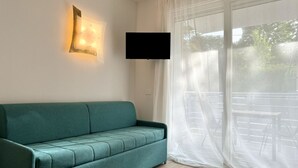 A teal sofa in a room with a large window, a mounted flat-screen TV, and a wall-mounted light fixture. - Residence Eva 6 - Carraro Immobiliare Jesolo (Cavallino-Treporti)