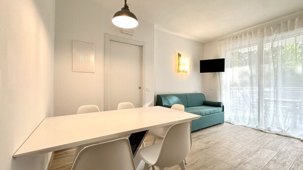 A modern living room with a white dining table, white chairs, a teal sofa, a flat-screen TV, and large windows with sheer curtains.