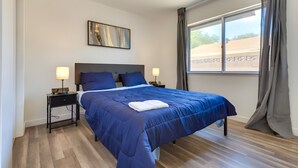 A neatly made bed with a blue comforter and pillows, two bedside tables with lamps, a wall-mounted abstract painting, and a large window with curtains. - Peaceful & Modern Ridgecrest Retreat: Spacious 4-Bed Home w/ Private Yard (Ridgecrest)
