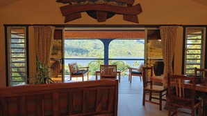 Ruime kamer met een groot raam met uitzicht op een meer en heuvels. Er staat een houten bank en een eettafel met stoelen.