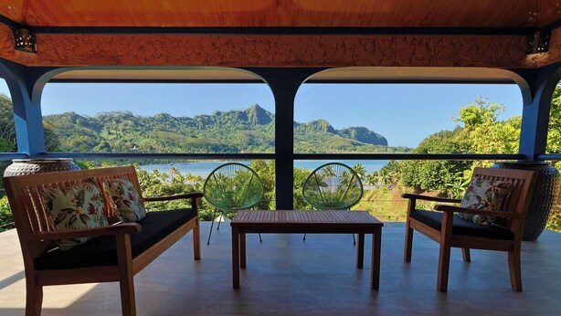 Een overdekt terras met twee houten stoelen en een tafel, met uitzicht op een mooi landschap met heuvels en water.