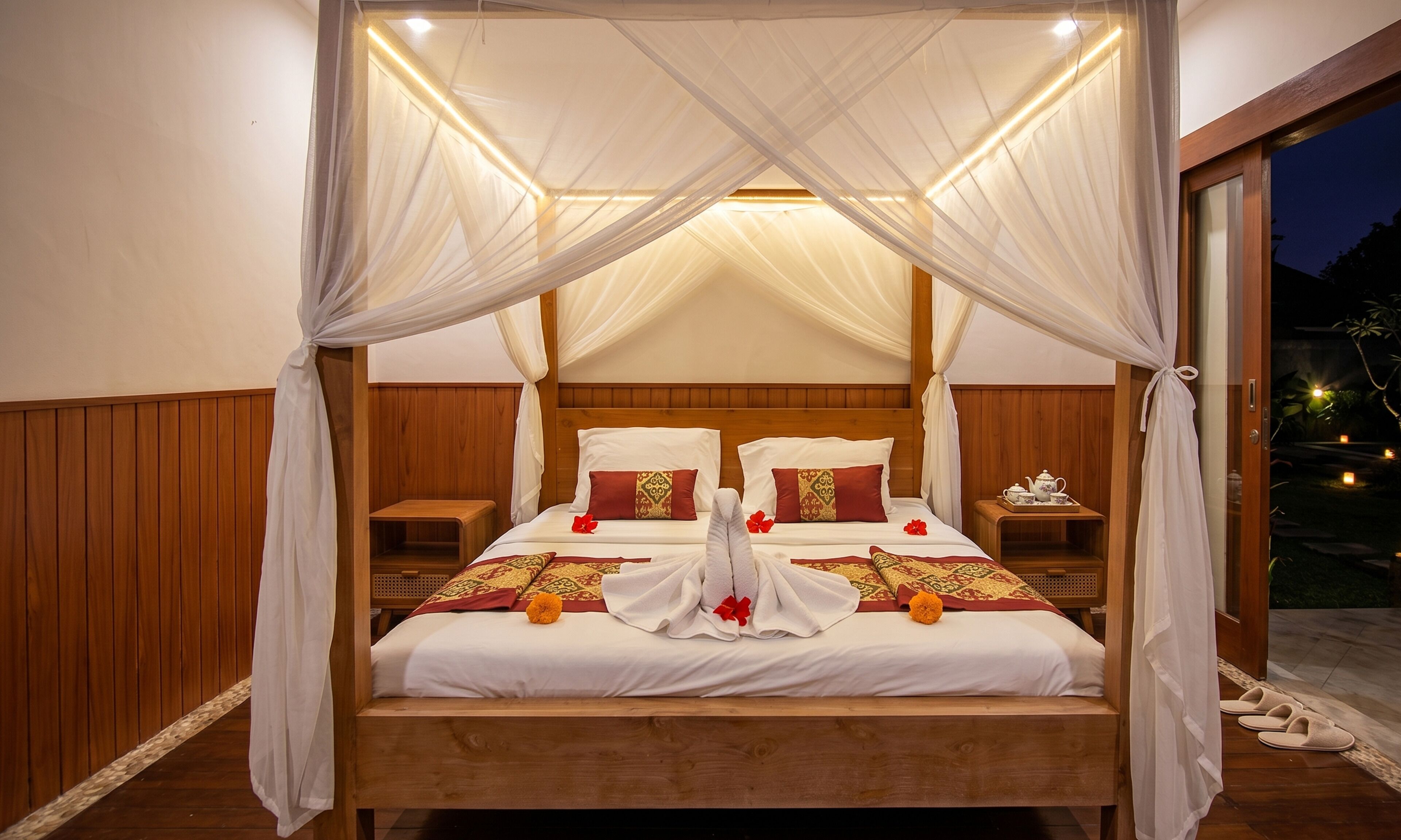 A four-poster canopy bed with white linens, two pillows with red and gold patterns, and a nightstand with a teapot and tray.