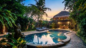 A swimming pool surrounded by lush greenery, a thatched-roof structure, and a seating area with lounge chairs. - Villa Sujati (Ubud)