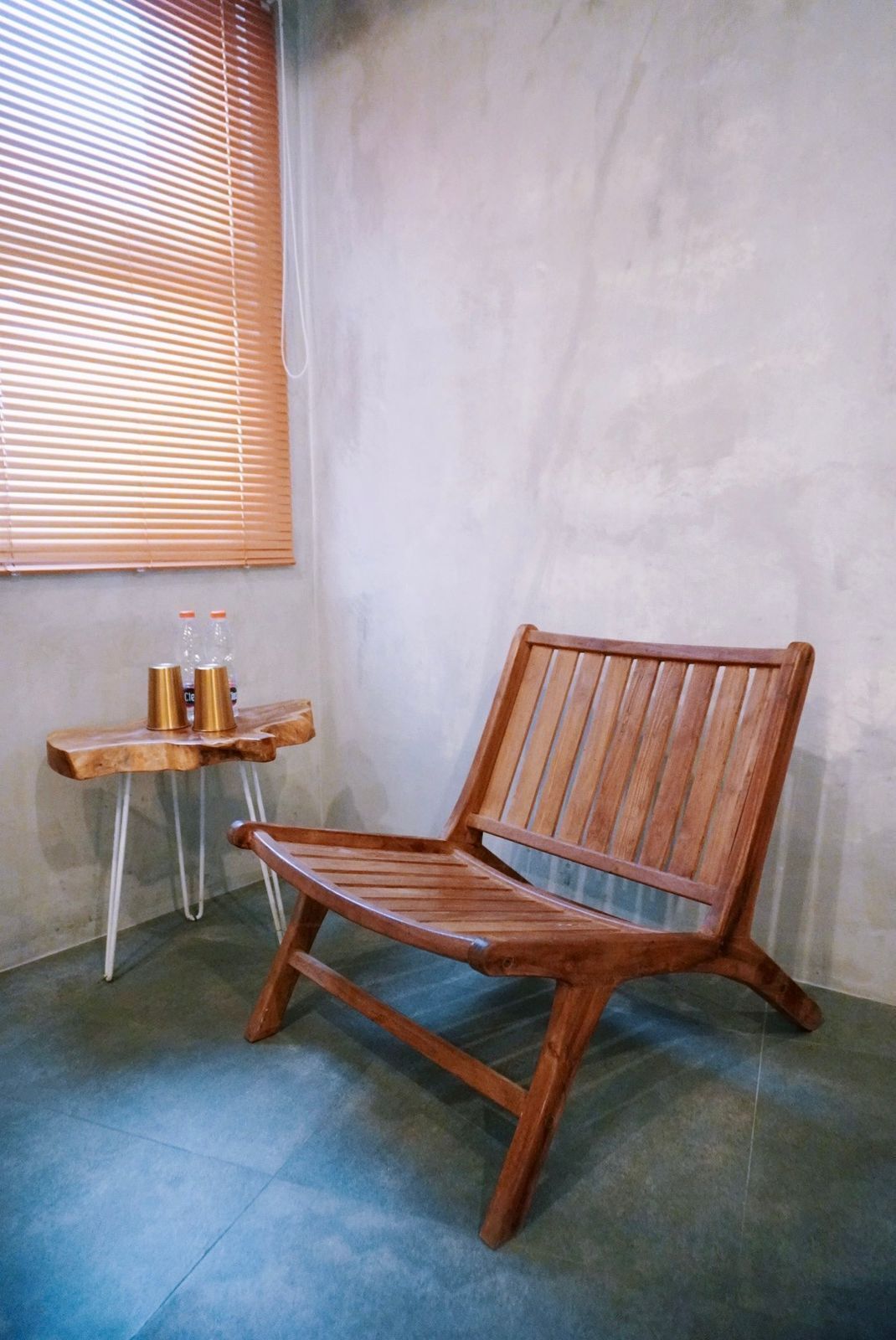 Een houten stoel met een latten rugleuning en armleuningen, geplaatst in een hoek van een kamer met een betonnen vloer en een raam met zonwering.
