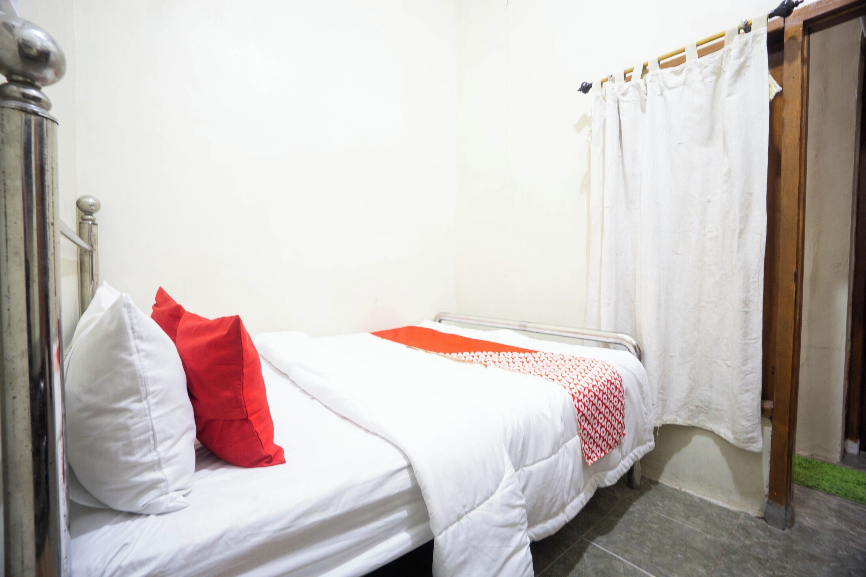 A small, single-bed room with a white bedspread, a red pillow, and a white curtain.