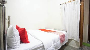 A small, single-bed room with a white bedspread, a red pillow, and a white curtain.