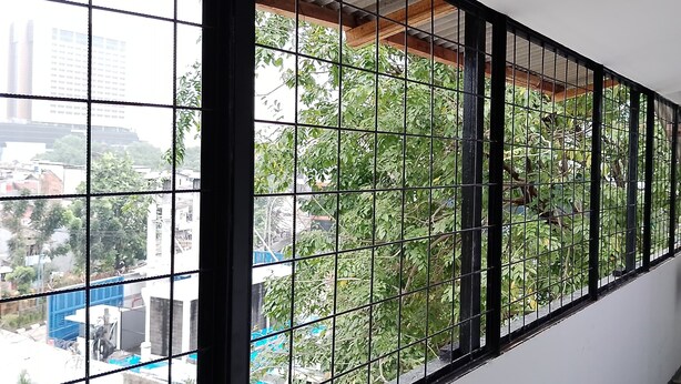 A view through a window with a metal grid, overlooking a building and greenery. - GoHome Harmoni Apartment (Jakarta)