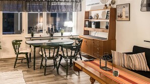 Een eetgedeelte met een ronde tafel en stoelen, een keuken met een houten kast, en een zithoek met een bank en een gestreept kussen.