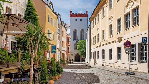 A cobblestone street lined with traditional buildings, a tower with a red roof, and outdoor seating with potted plants.