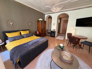A bedroom with a bed, bedside tables, a TV, a dining area with chairs, and a hallway.