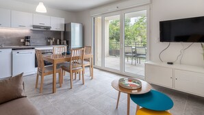 A modern kitchen with a dining area, a TV mounted on the wall, and a balcony with a view of greenery.