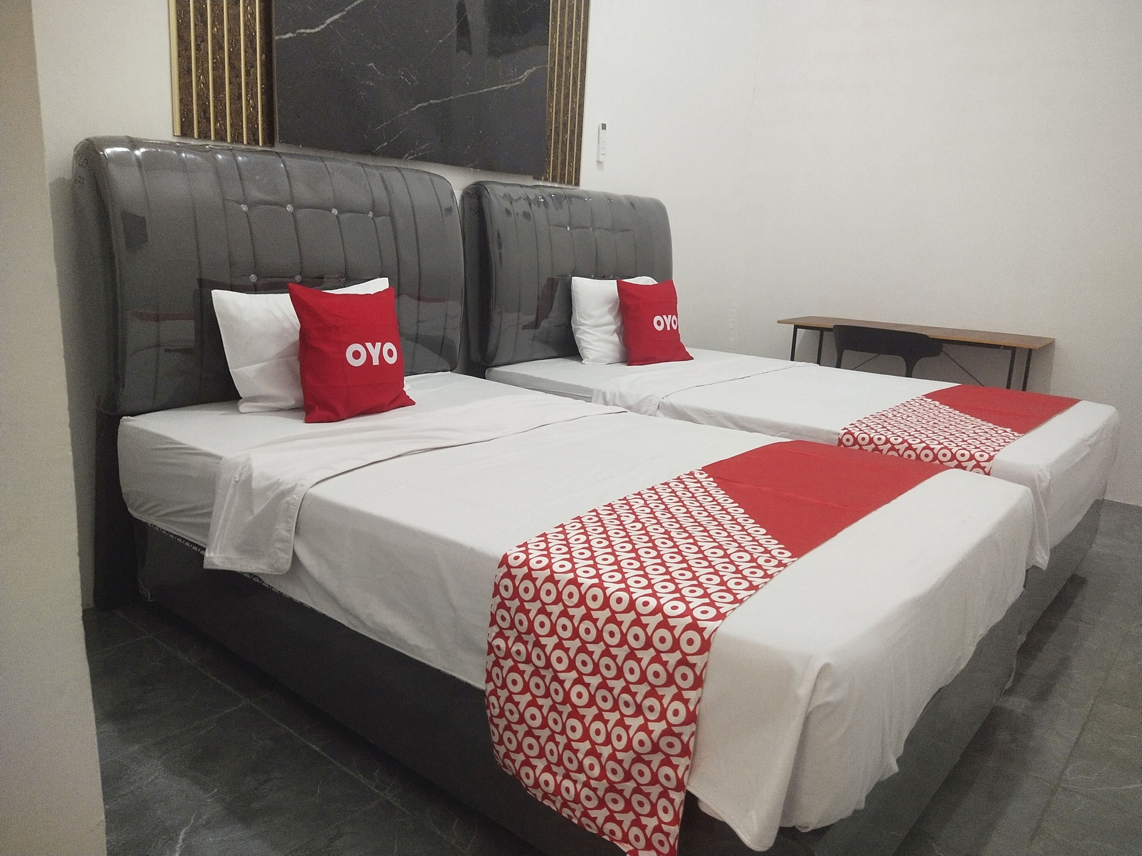 A hotel room with two beds, each with a white bedspread and a red patterned blanket. There are two red pillows with the word "OYO" on them. The room has a dark headboard and a small wooden table beside the beds.