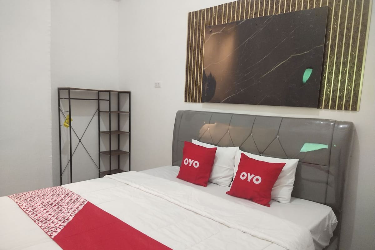 Una cama bien hecha con una colcha blanca y roja y dos almohadas con el texto “OYO”, un cabecero tapizado gris y una obra de arte con marco negro y dorado encima.