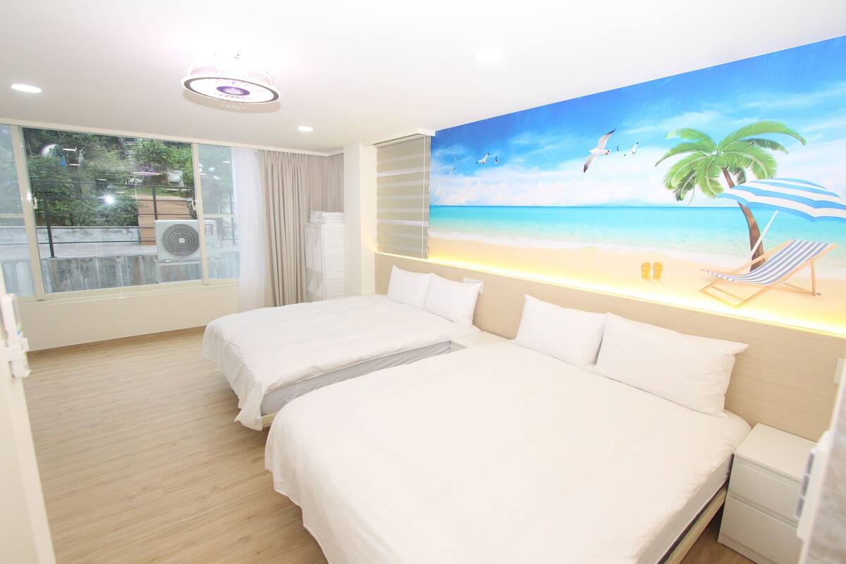 Une chambre d’hôtel avec deux lits, une grande fenêtre donnant sur la mer et une fresque murale sur le thème de la plage.