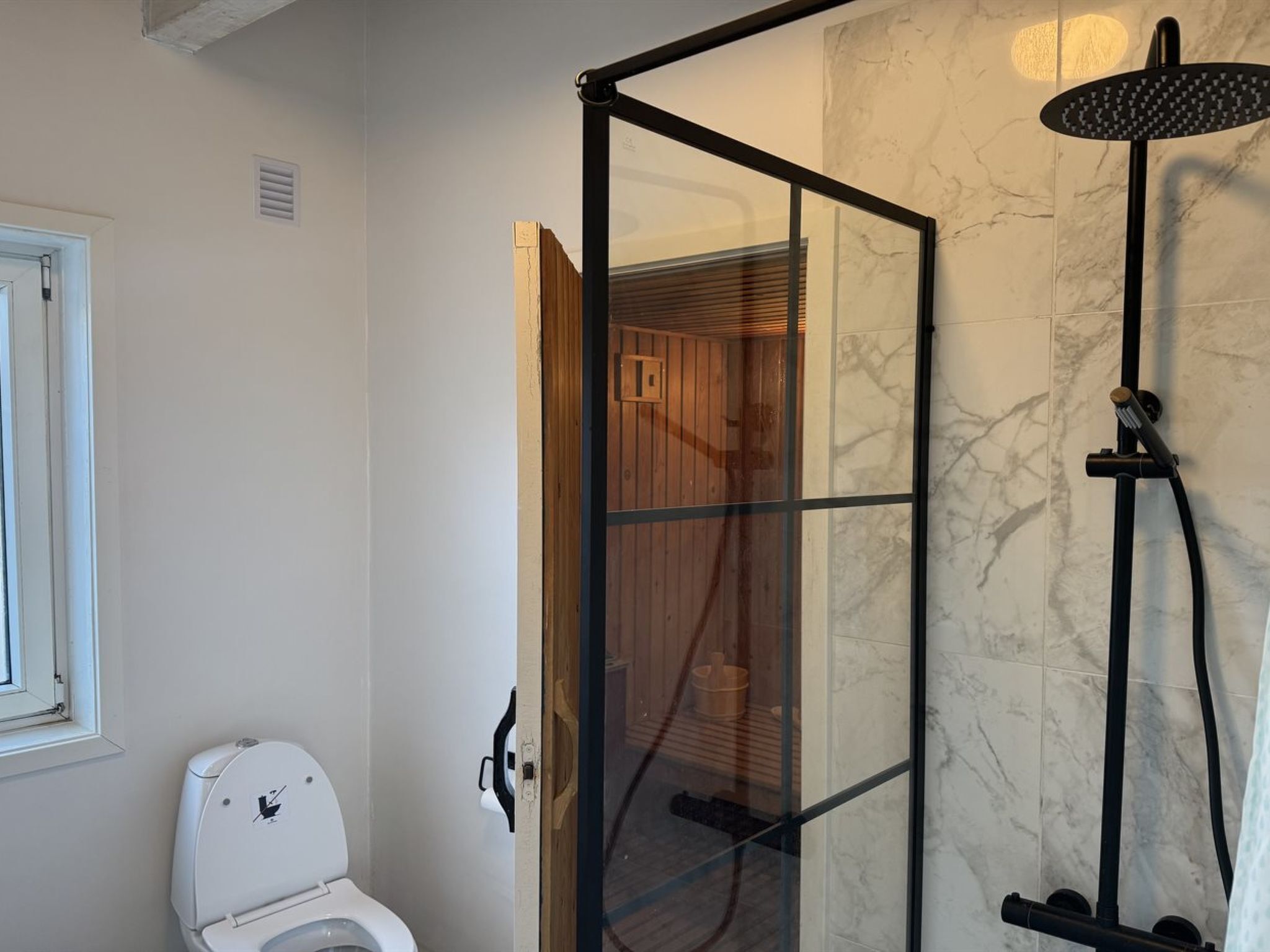 Et badeværelse med en glasafskærmet brusekabine, et hvidt toilet og en marmorvæg.